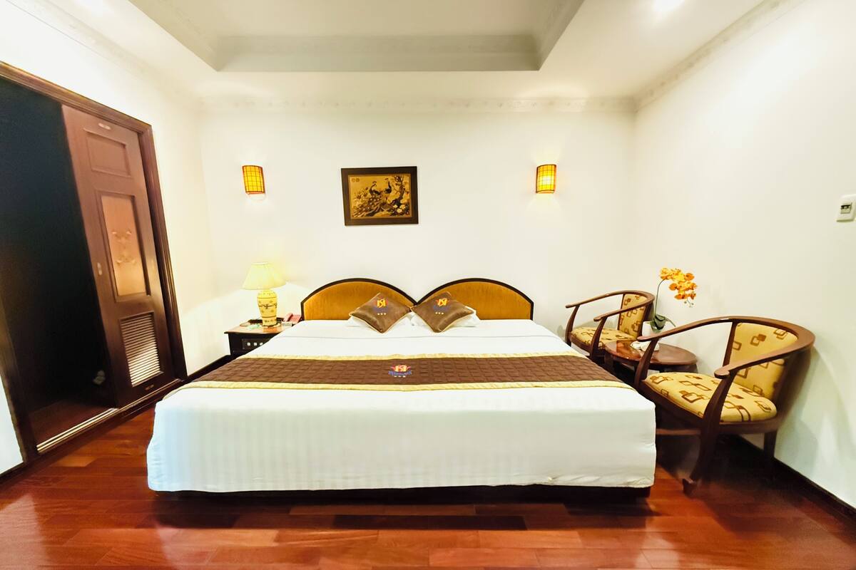 Habitación de hotel con una cama grande, cabecera de madera, dos lámparas de noche, un cuadro en la pared y una silla con una mesita.