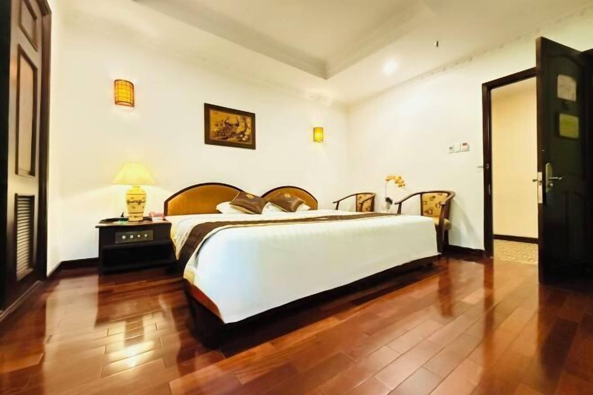 Habitación de hotel con piso de madera, una cama con cubrecama blanco, una mesita de noche con lámpara y una puerta a la derecha.
