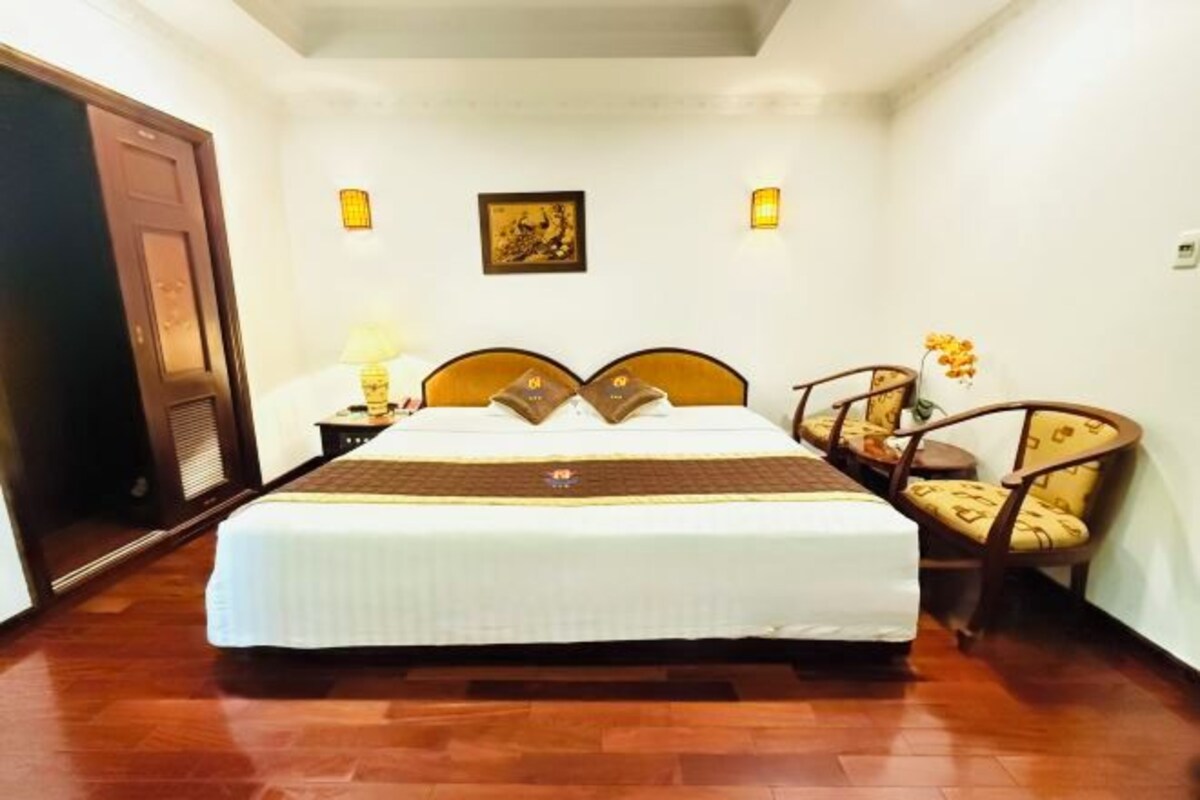 Una habitación de hotel con cama, mesita de noche, lámpara, silla e imagen colgada en la pared.