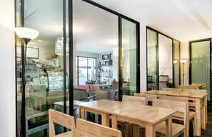 A modern interior with wooden tables and chairs, large windows, and a visible kitchen area.