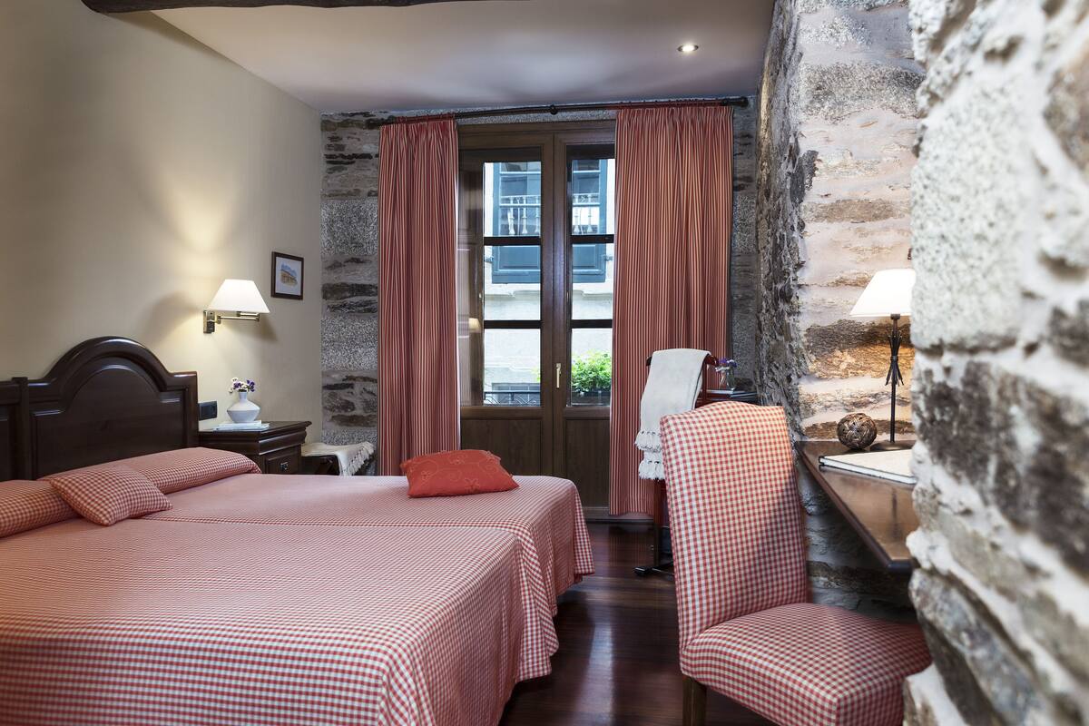 A hotel room with a checkered bedspread, a wooden headboard, a nightstand with a lamp, a chair, and a stone wall.