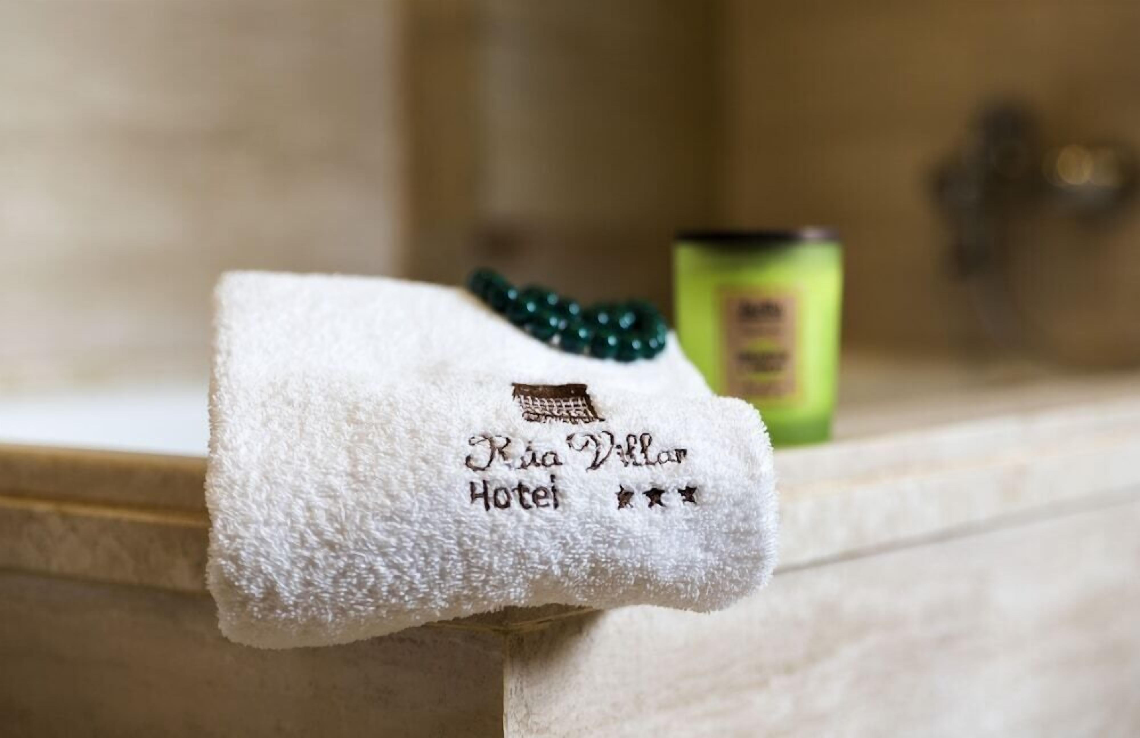 A white towel with 'Ria Villar Hotel' embroidered on it, a green bottle, and a green beaded bracelet on a bathroom counter.