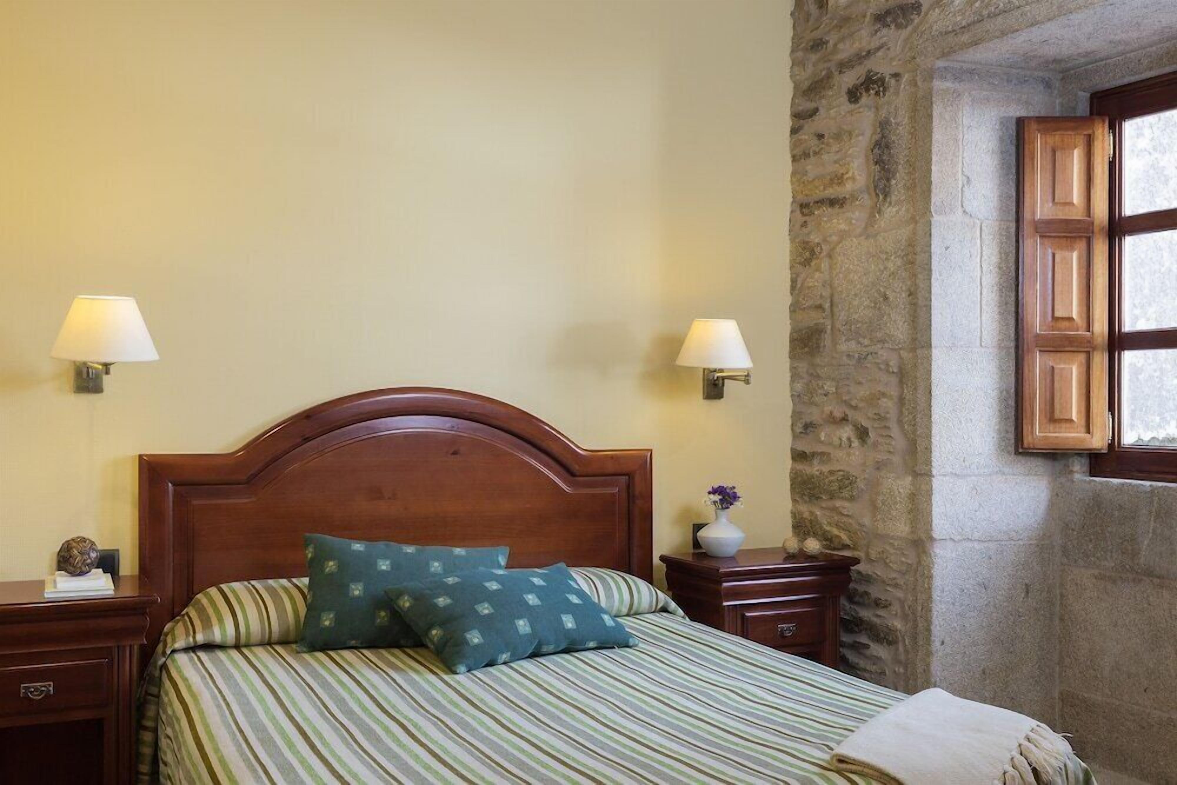 Un dormitori amb un panell de fusta al capçal, dues taules de nit amb làmpades, un llit amb roba de llit a ratlles i una paret de pedra.