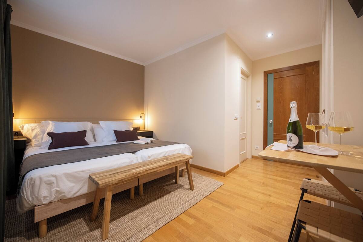 Ein Schlafzimmer mit einem Bett, Nachttischen, einer Bank und einem kleinen Tisch mit Flasche und Gläsern.
