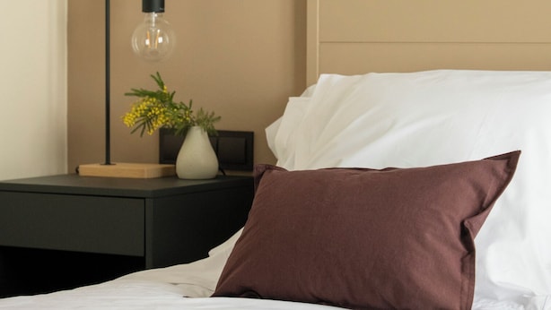 Ein ordentlich bezogenes Bett mit weißen und braunen Kissen, ein Nachttisch mit Lampe und Blumenvase sowie ein beigefarbener Kopfteil.