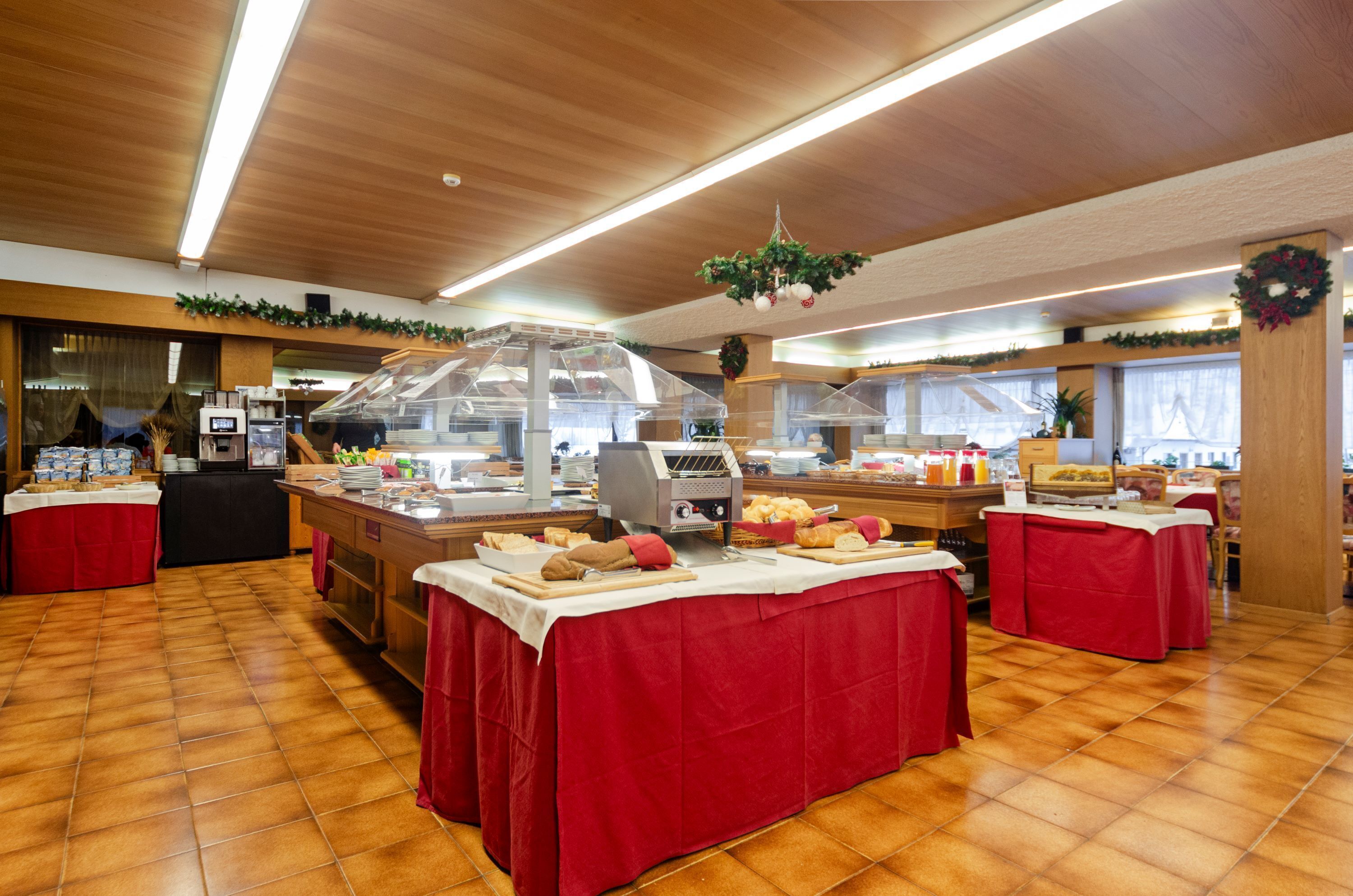 A buffet setup with multiple tables covered in red tablecloths, a self-service food station, and a wooden ceiling with hanging greenery.