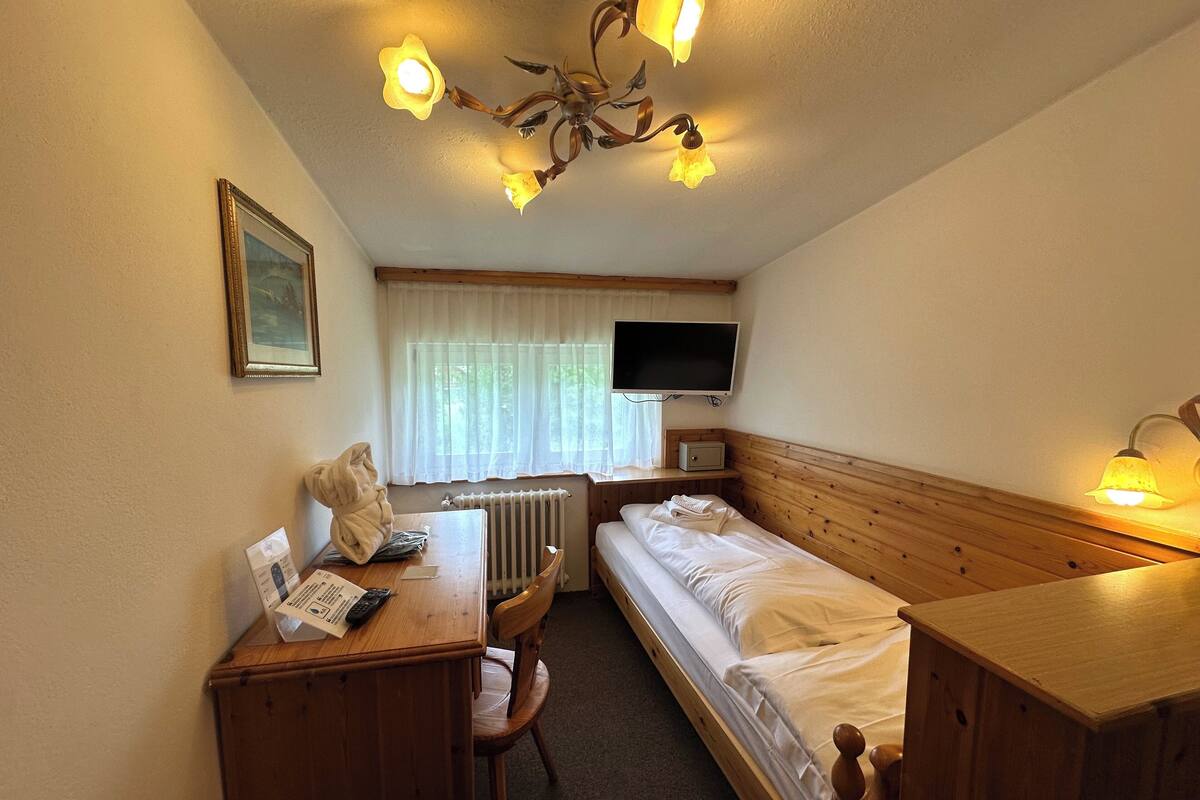 A small, cozy hotel room with a wooden bed, a desk with a telephone and papers, a chair, a TV mounted on the wall, and a ceiling light fixture.