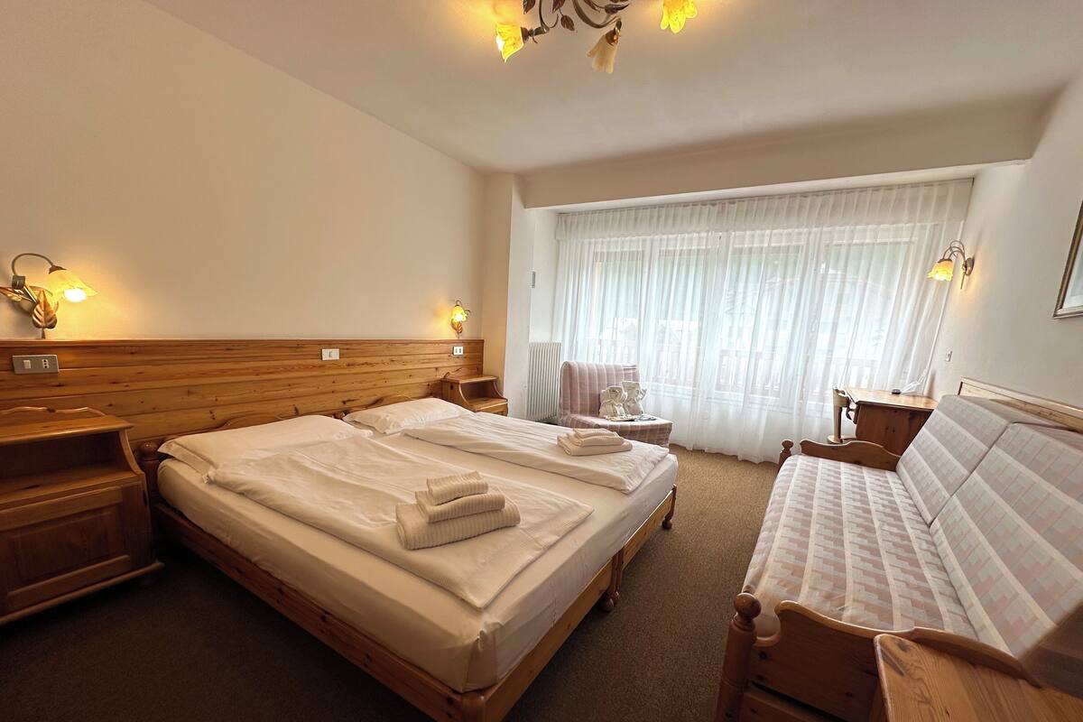 A hotel room with two beds, a wooden headboard, bedside lamps, a small table, and a window with sheer curtains.