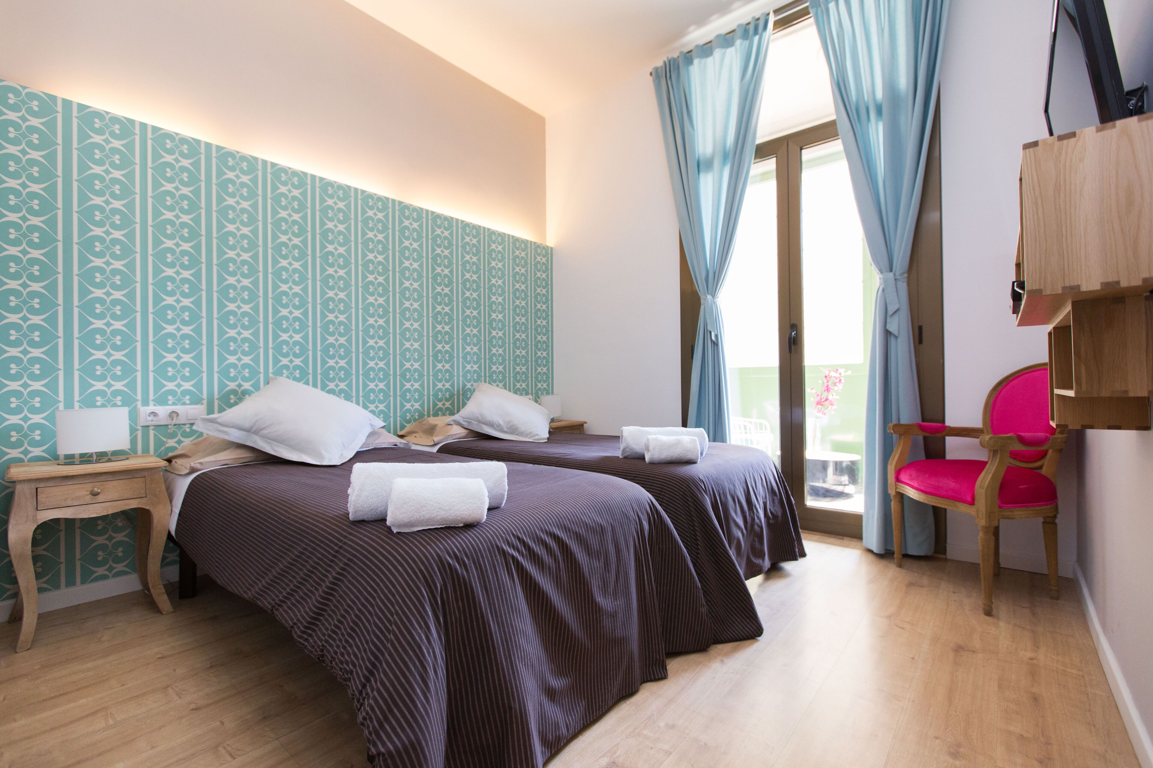 Una habitación de hotel con dos camas, una mesita de noche de madera, una silla y un ventanal con cortinas.