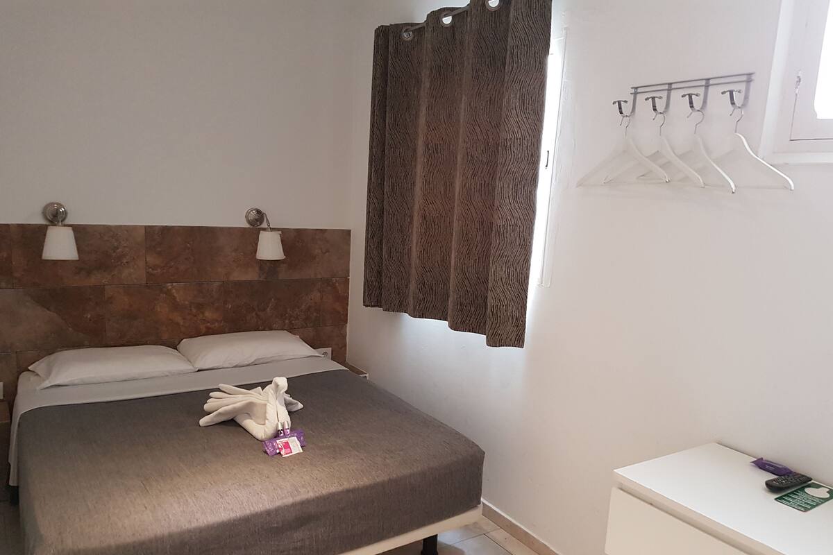 Una cama bien tendida con una colcha gris, dos almohadas y una toalla sobre ella. Se observa una mesita de noche blanca con un control remoto y una tarjeta verde. Además, un cabecero de madera y una lámpara de pared.