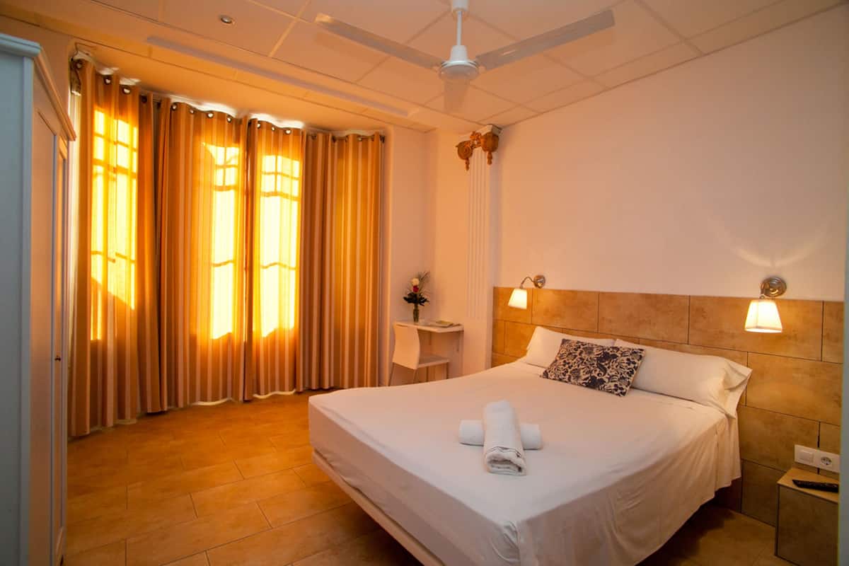 Habitación de hotel con una cama grande, una mesita de noche, un ventilador de techo y una ventana con cortinas.