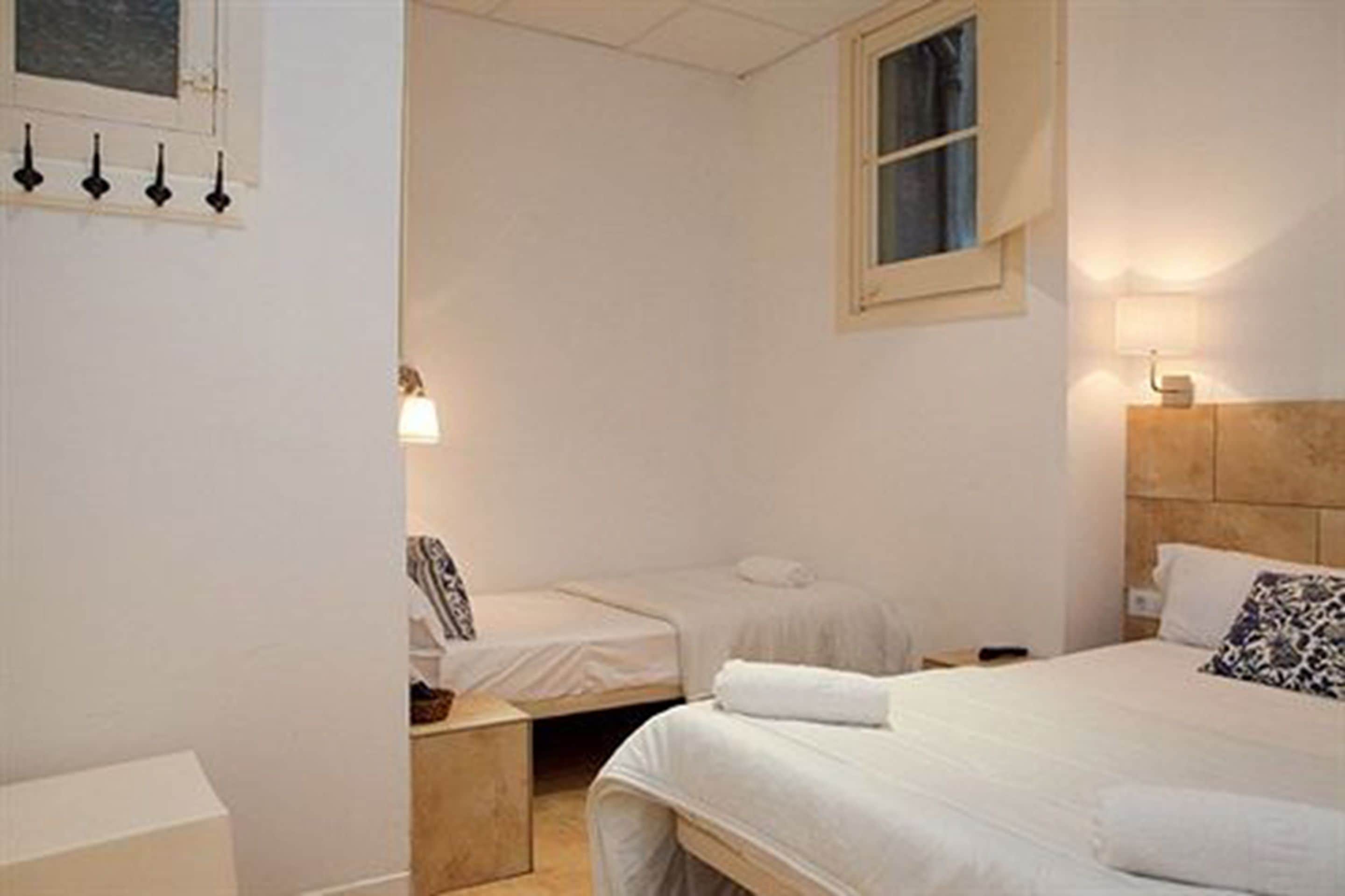 A hotel room with two single beds, a wooden headboard, a bedside table, a nightstand, a lamp, and a window with curtains.