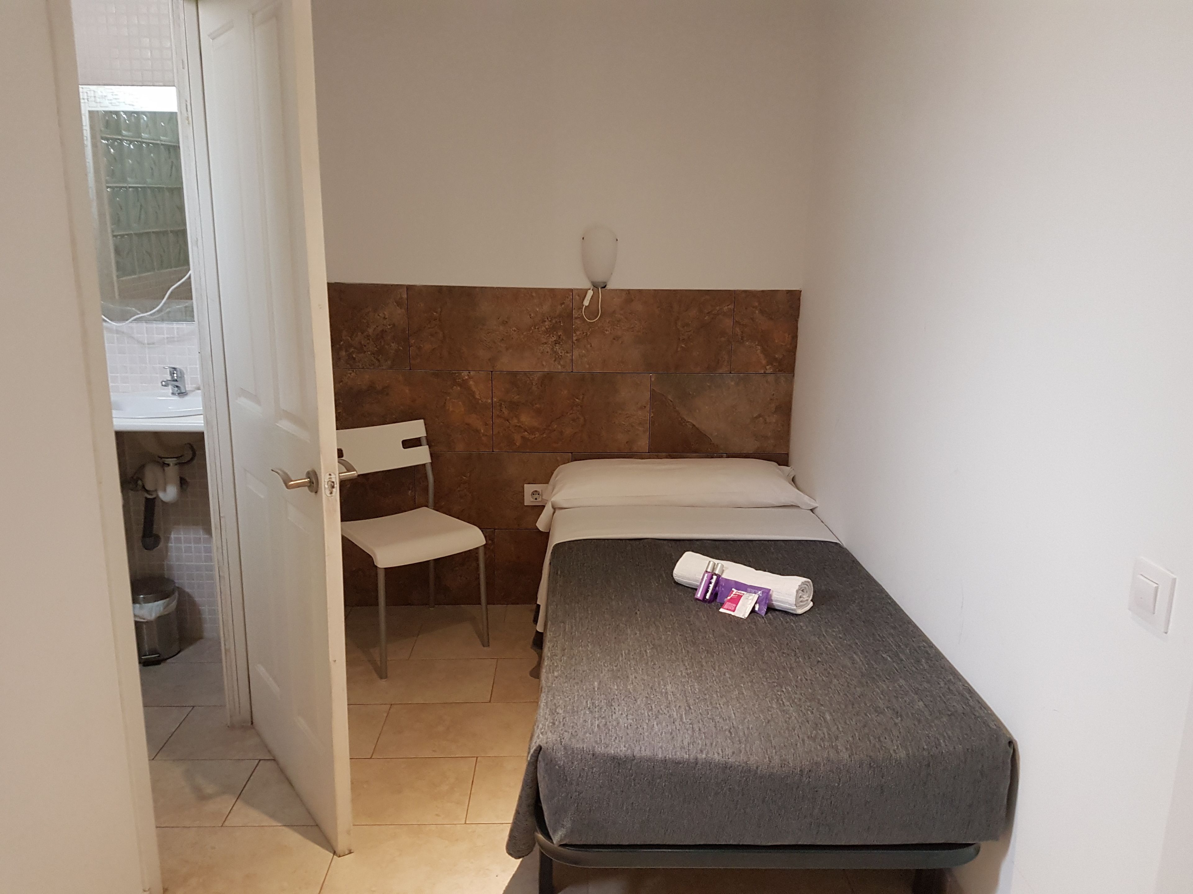 A small, compact room with a single bed, a chair, a small table, and a wall-mounted light fixture.