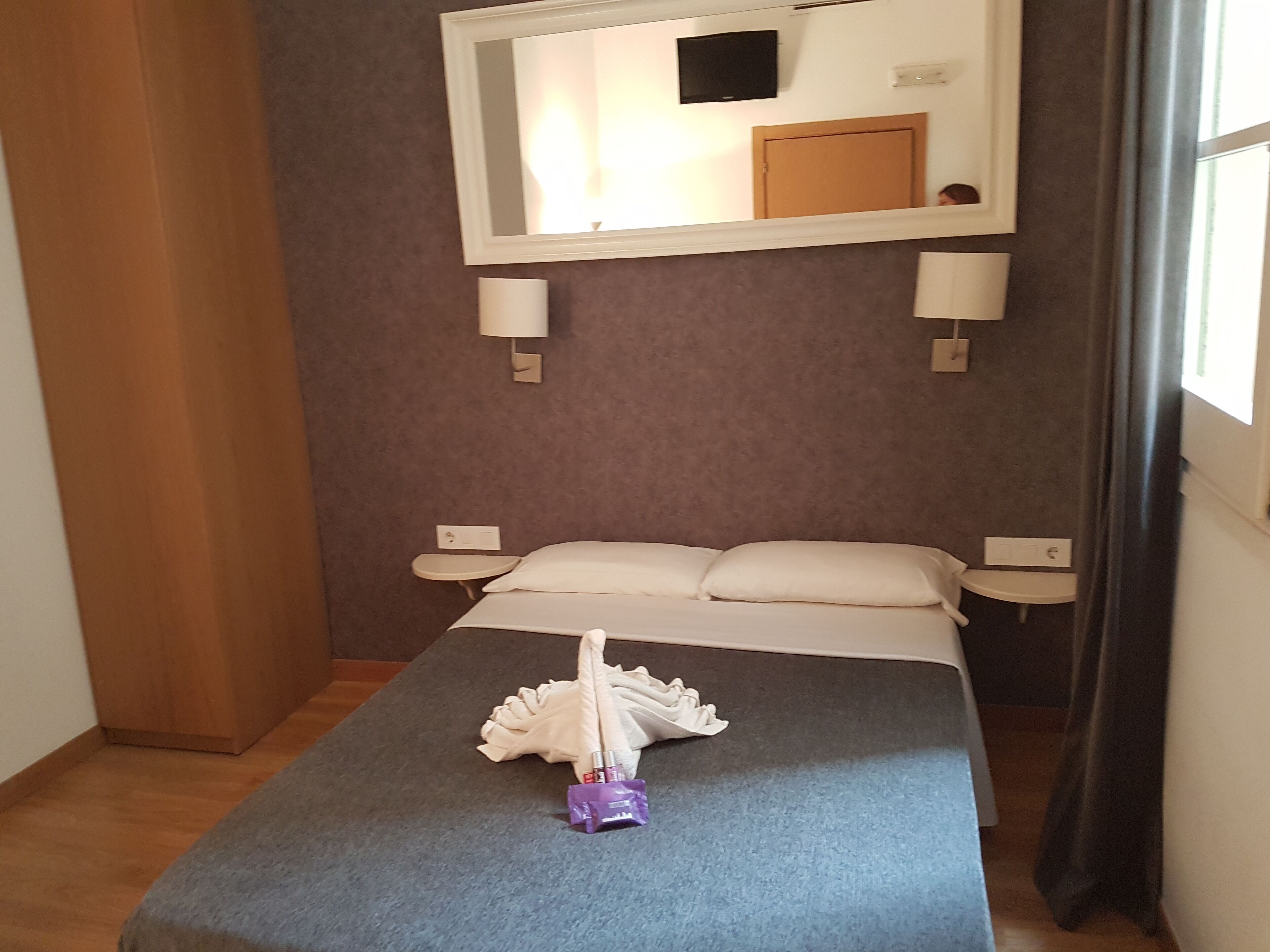 A hotel room with a bed, bedside tables, a mirror, and a window with curtains.