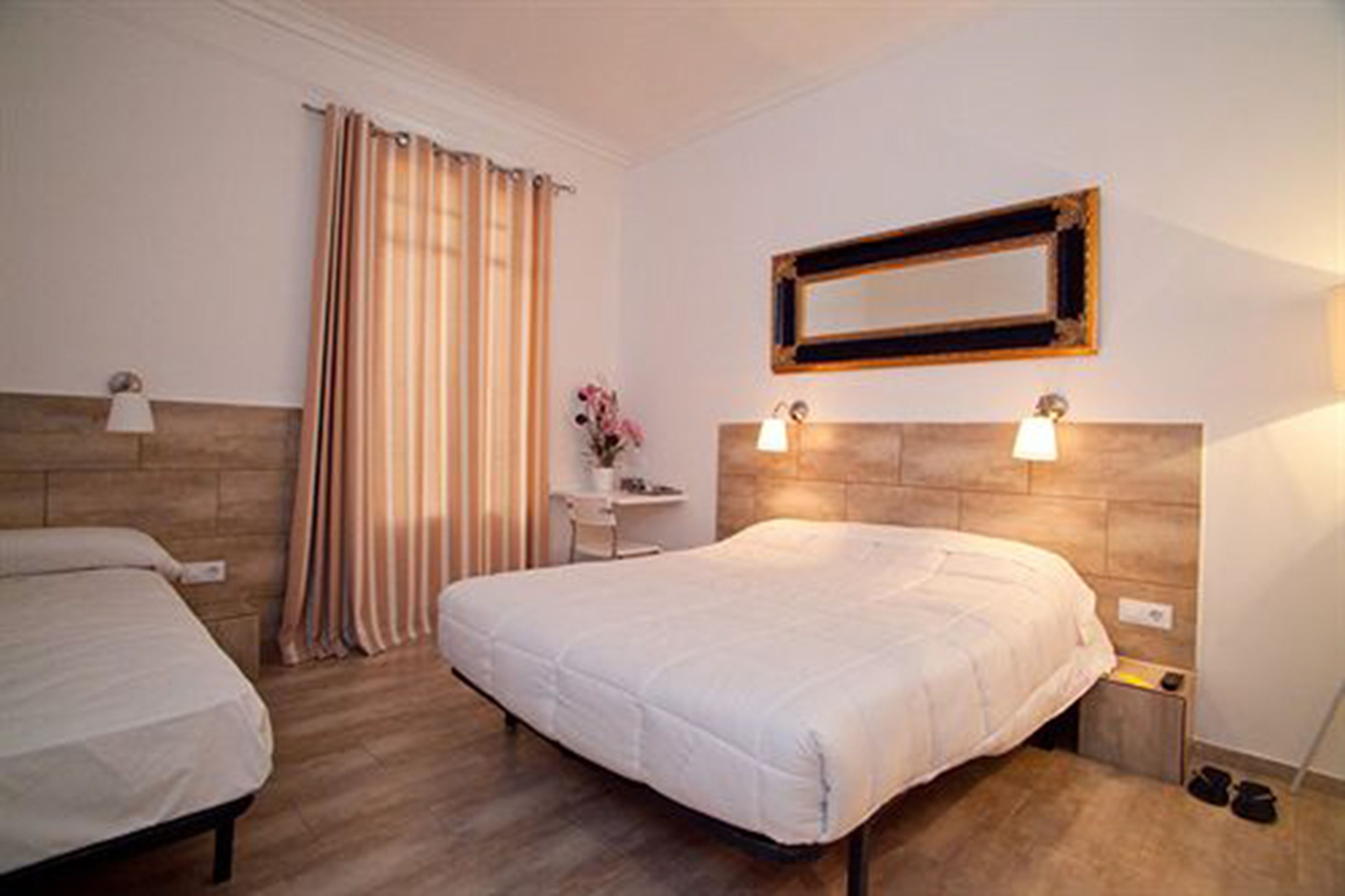 A hotel room with two beds, a wooden headboard, a small desk with a chair, a mirror, and a pair of shoes on the floor.