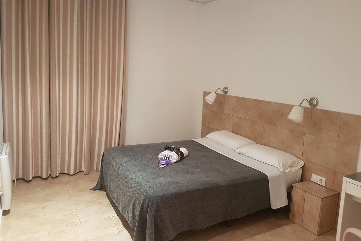 Una cama bien hecha con colcha gris y almohadas blancas, una mesita de noche con una lámpara y una ventana con cortinas color beige.