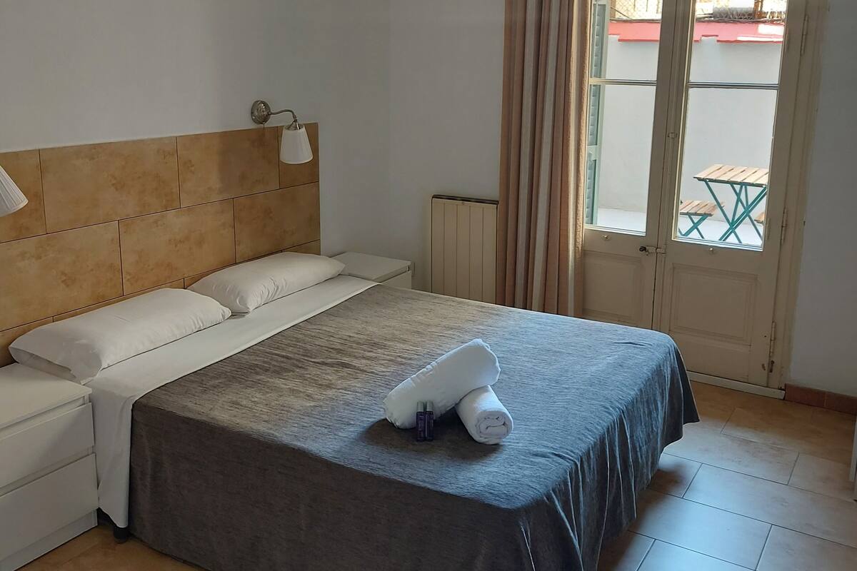 Habitación de hotel ordenada con cama, mesita de noche, lámpara y ventana con cortinas.