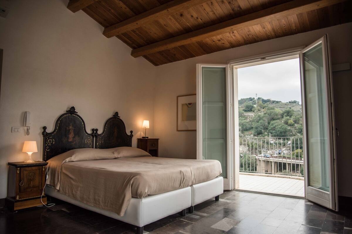A bedroom with a bed, bedside lamps, a wooden ceiling, and a balcony with a view of greenery.