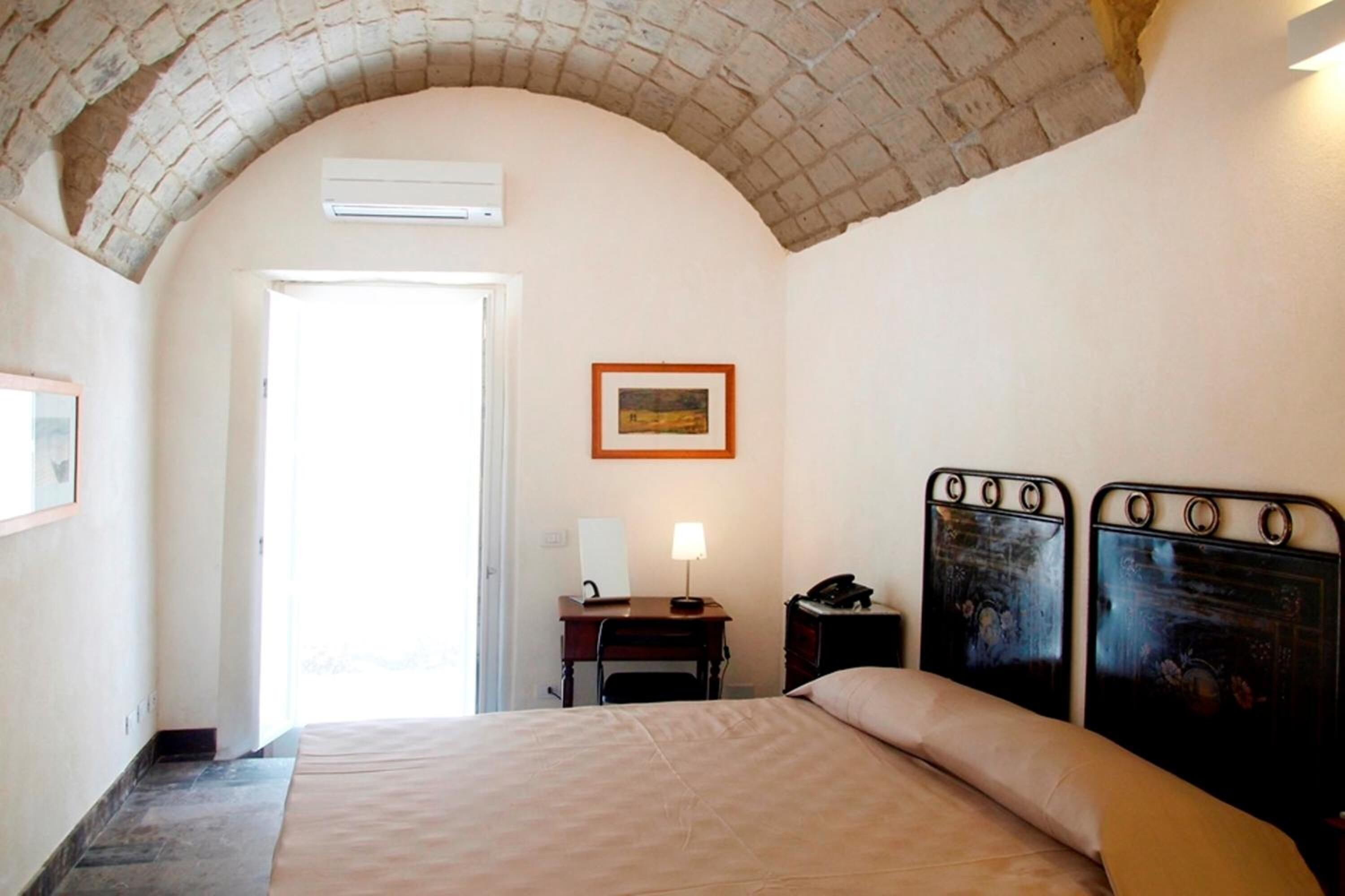 A bedroom with a bed, a desk with a lamp, a telephone, and a framed picture on the wall.