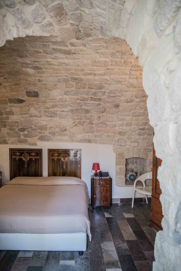 Un dormitorio con pared de piedra, una cama, una mesita de noche, una lámpara roja y una silla.