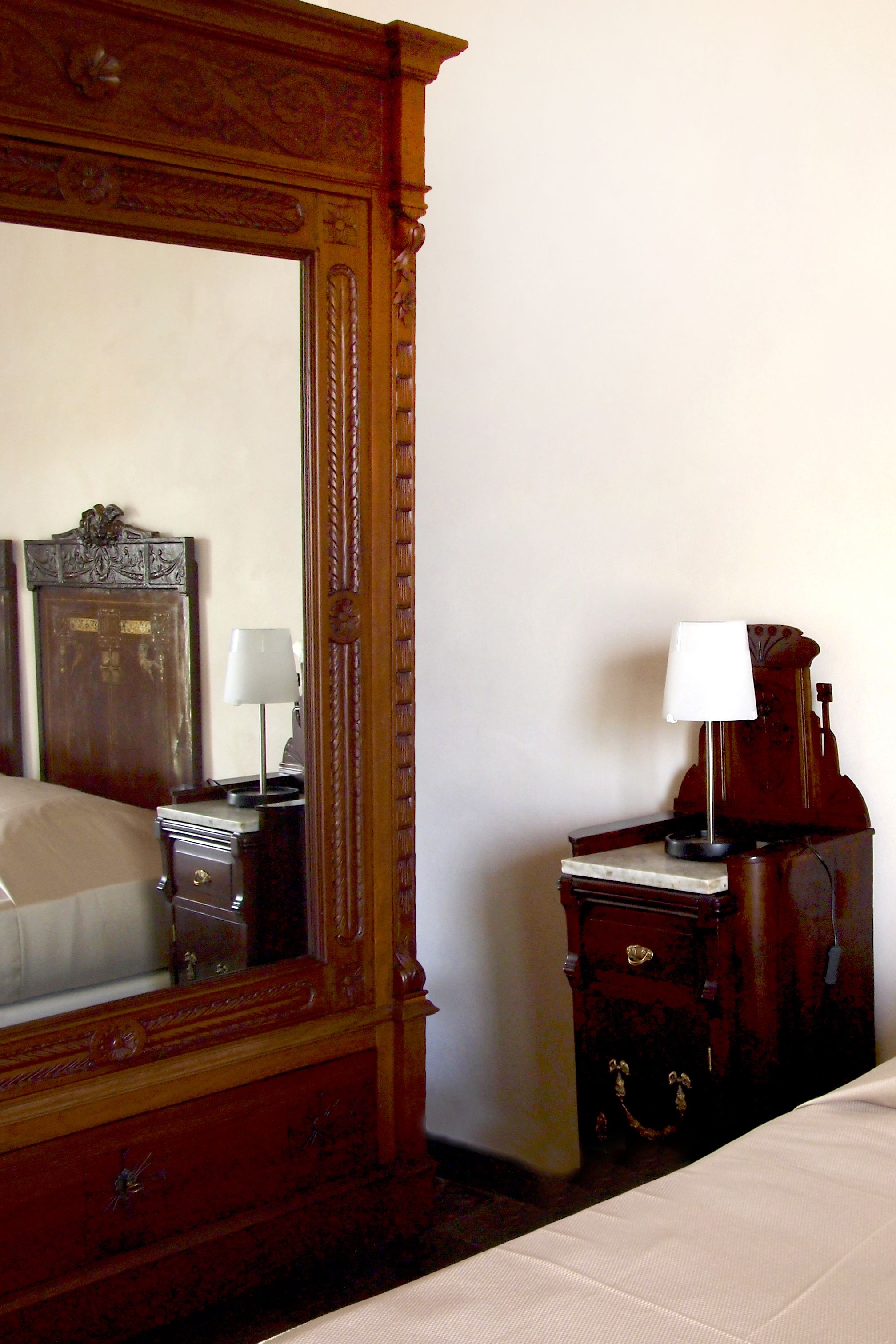 A hotel room with a large ornate mirror, a wooden nightstand with a lamp, and a bed with a white bedspread.