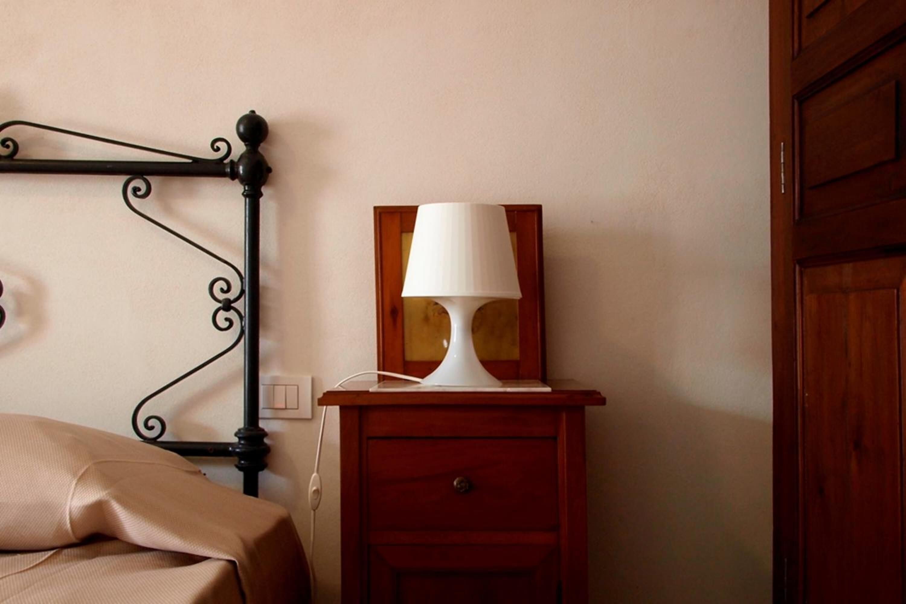 A bed with a beige cover, a bedside table with a white lamp, and a wooden nightstand with a drawer.