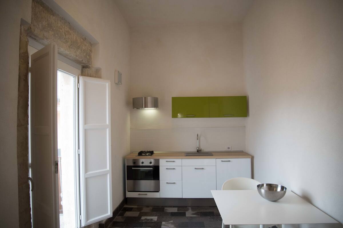 A compact kitchen with white cabinets, a green backsplash, a stainless steel sink, and a white dining table with a metal bowl.