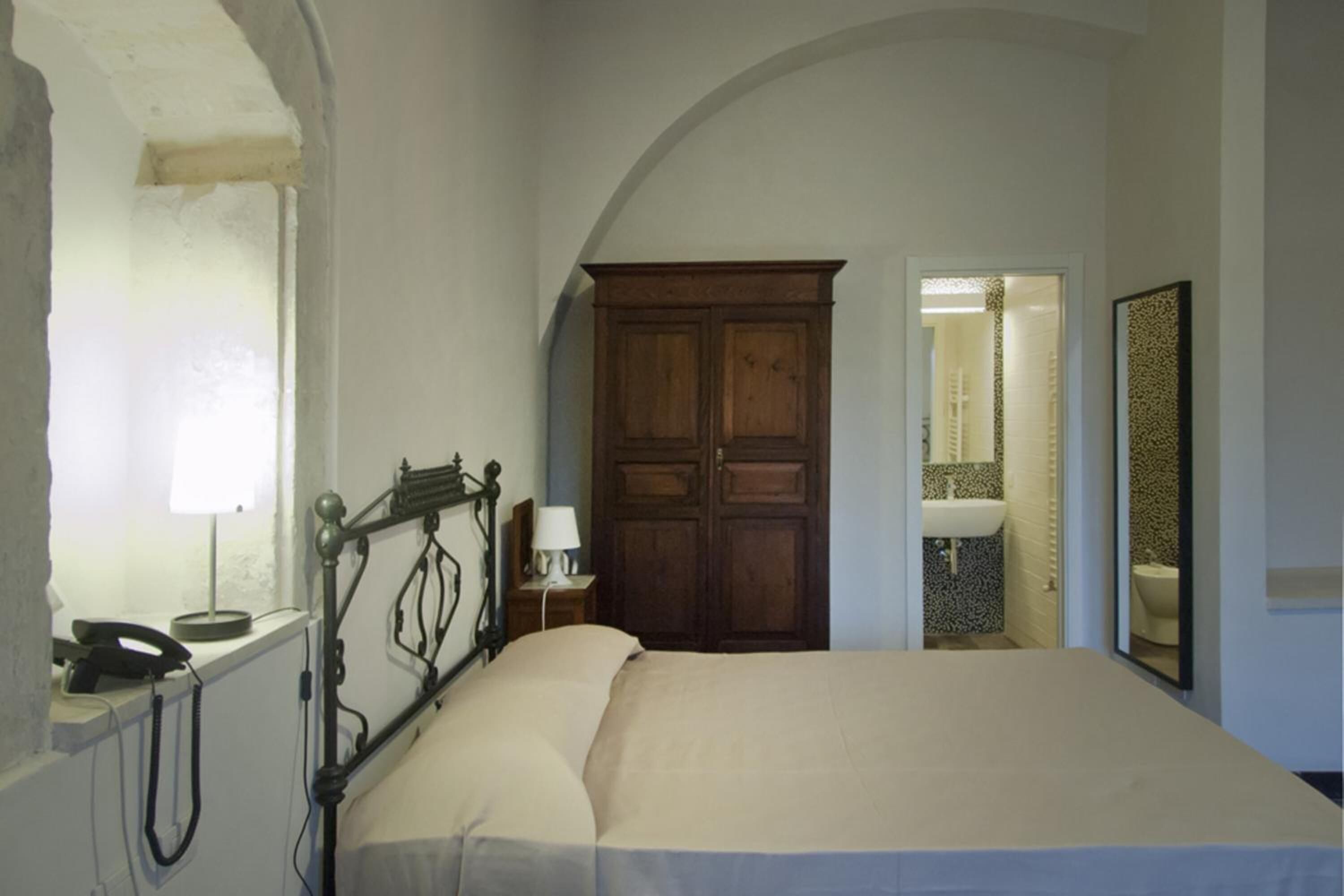 Un dormitorio con una cama grande, un armario de madera, una mesita de noche con lámpara, y un baño visible a través de un arco.