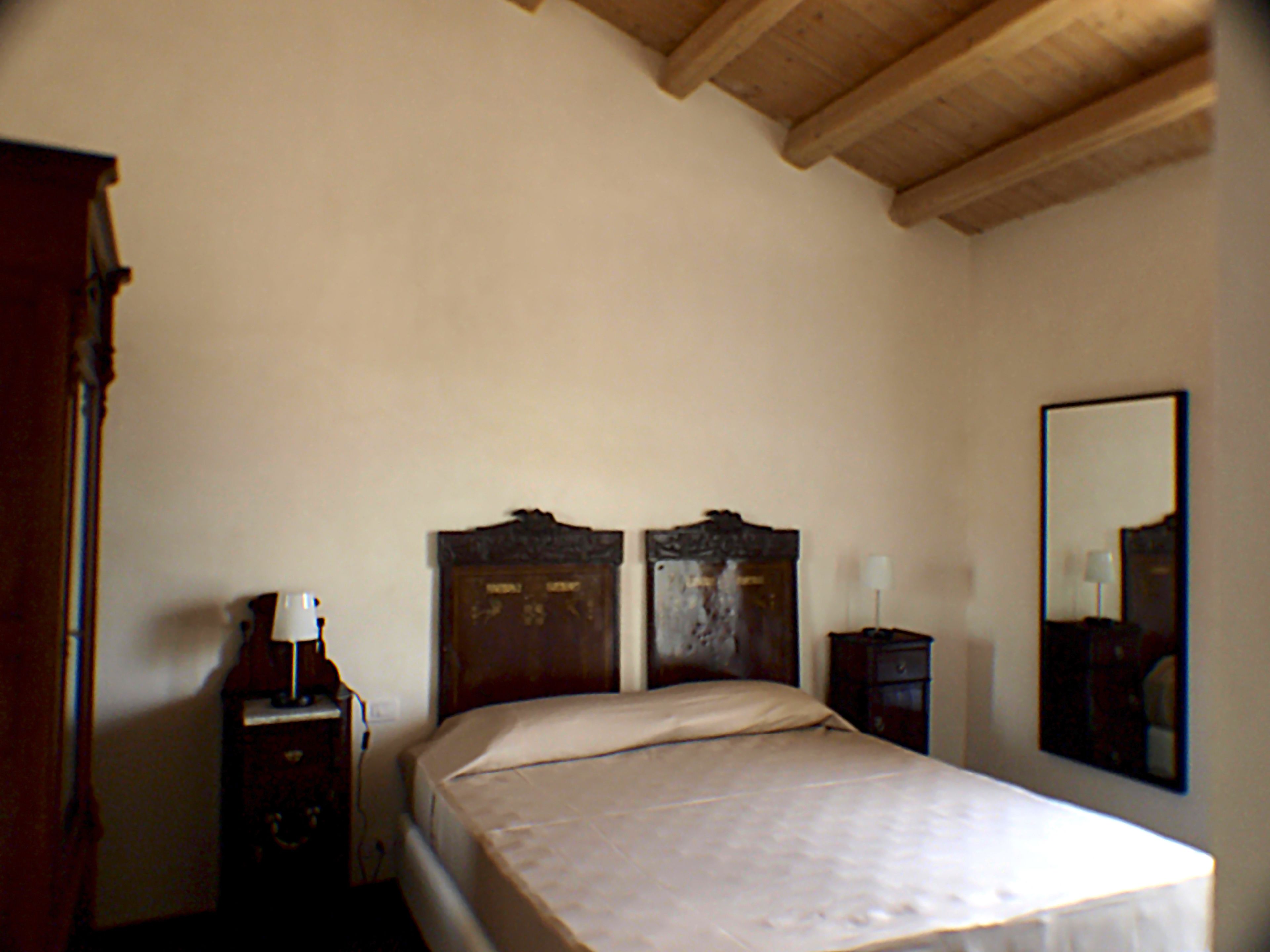 A bedroom with a wooden ceiling, a bed with a white mattress, a wooden nightstand with a lamp, and a wooden dresser.