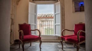 A room with two red upholstered chairs facing a window with an open shutter, revealing a view of buildings with tiled roofs.