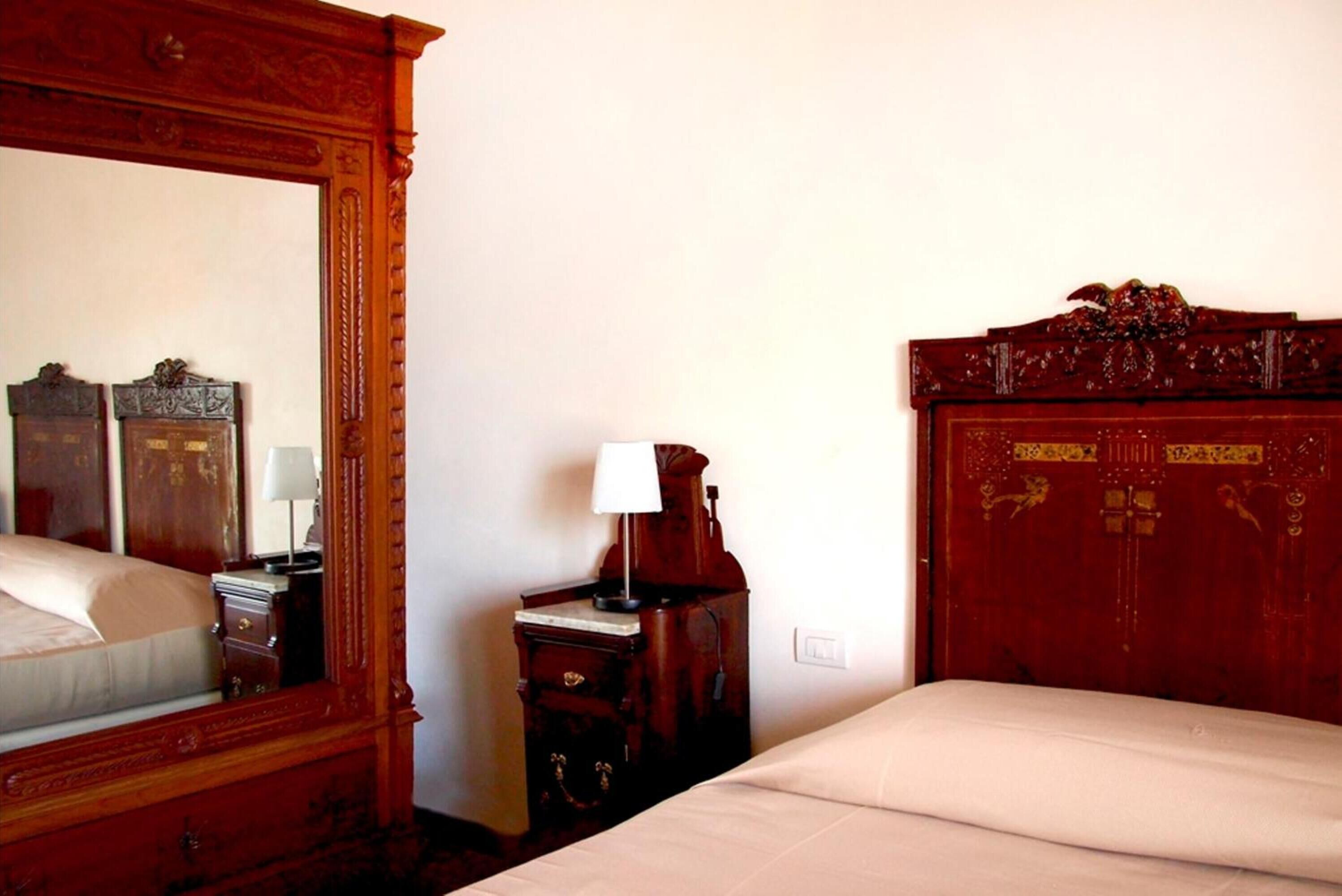 A hotel room with a large mirror, a wooden wardrobe, a bedside table with a lamp, and a bed with a white comforter.