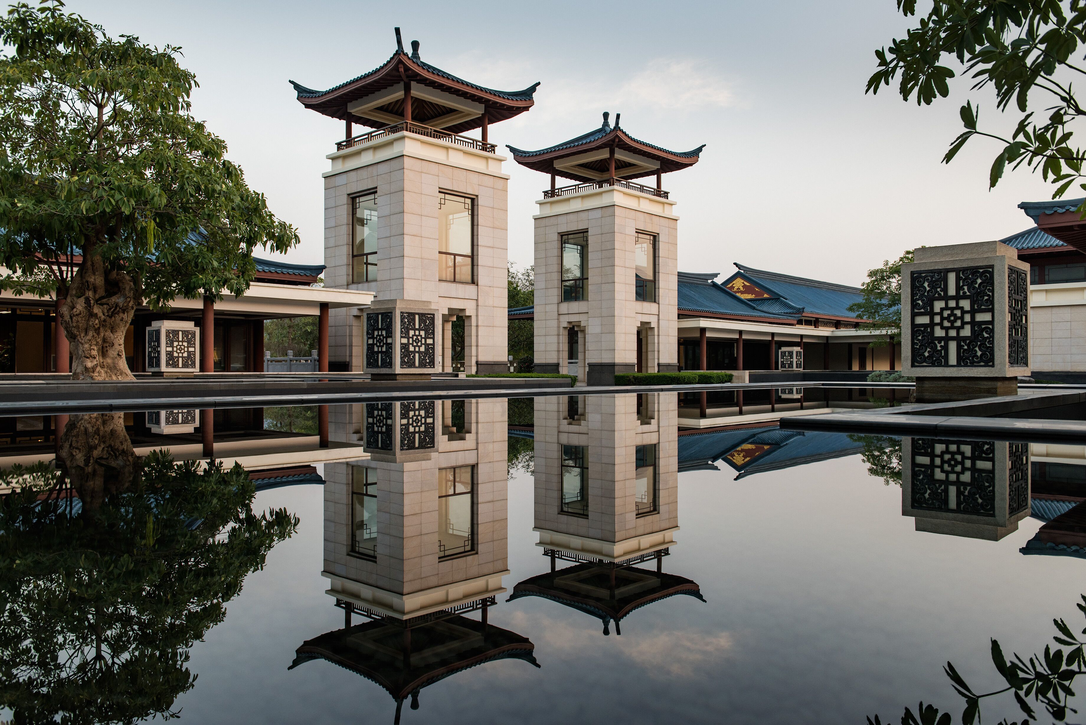 Un edificio tradicional con dos torres al estilo pagoda, una superficie acuática reflectante y un árbol en primer plano.