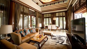 A spacious living room with a large sofa, a coffee table, and a television. The room has large windows with curtains, a chandelier, and a patterned carpet. - Imperial Springs (Guangzhou)