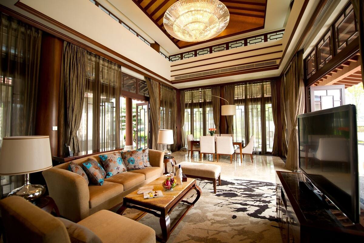 A spacious living room with a large sofa, a coffee table, and a television. The room has large windows with curtains, a chandelier, and a patterned carpet.