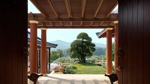 A traditional Japanese-style room with tatami flooring, wooden pillars, and a view of a landscaped garden with a large tree and mountains in the background. - Imperial Springs (Guangzhou)