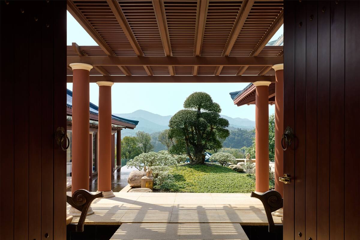 A traditional Japanese-style room with tatami flooring, wooden pillars, and a view of a landscaped garden with a large tree and mountains in the background.