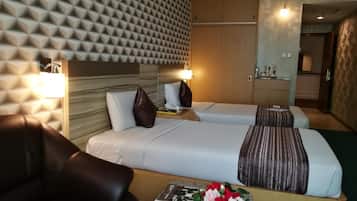 A hotel room with two beds, a sofa, a desk with a magazine and a flower arrangement, and a kitchenette in the background.