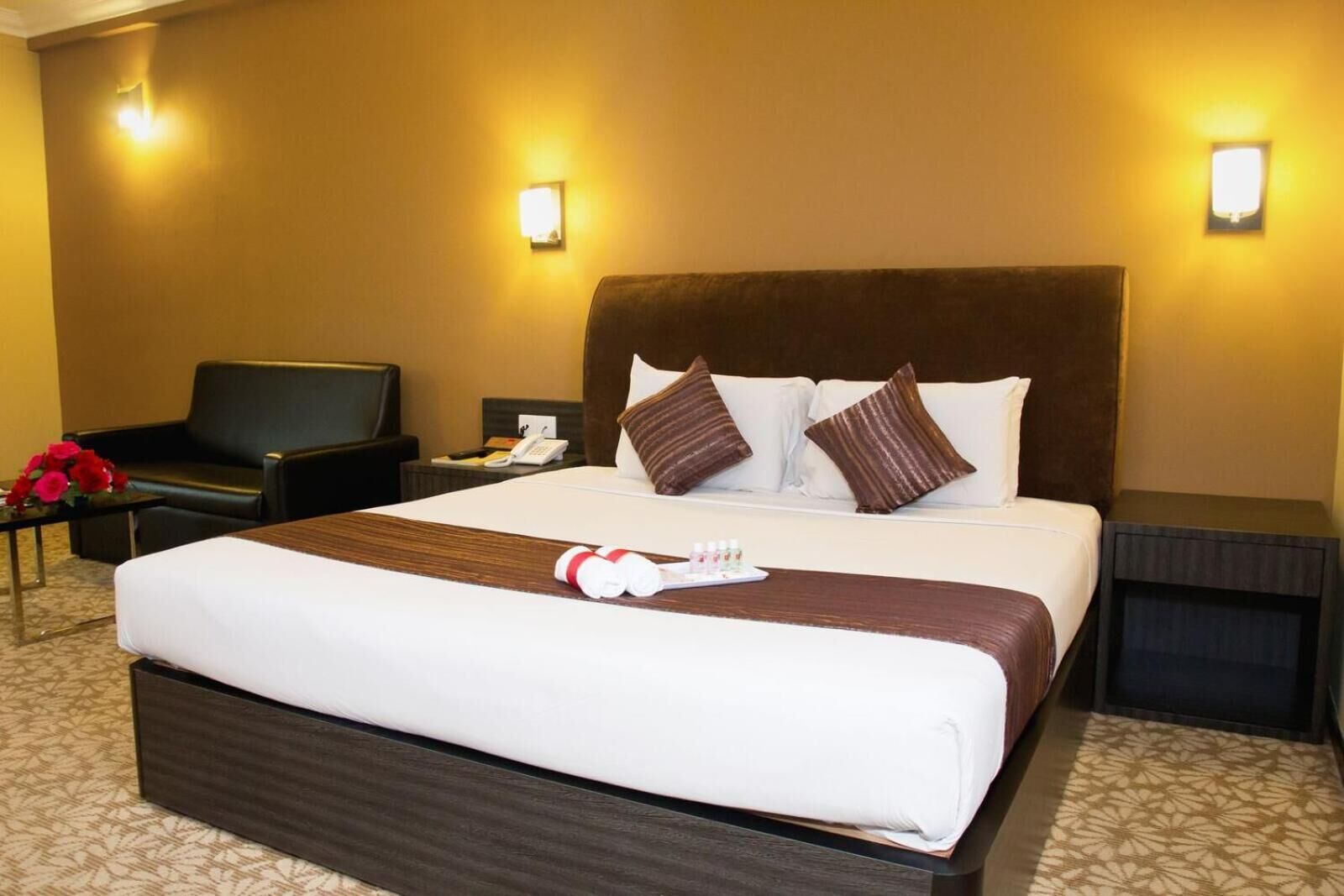 A hotel room with a large bed, a bedside table with a lamp, a chair, and a small table with a vase of flowers.