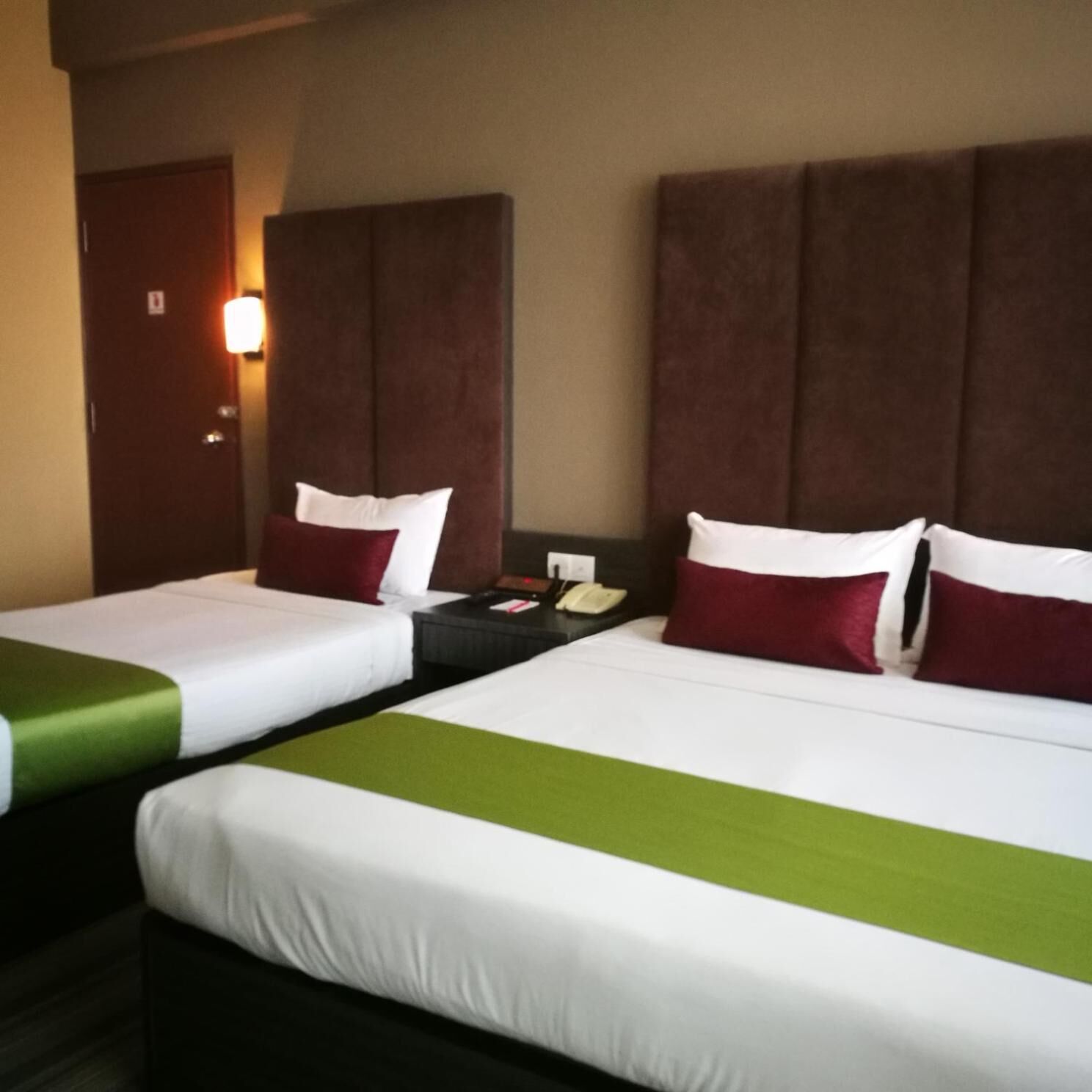 A hotel room with two beds, each with white and green bedding, and brown headboards. There is a nightstand with a phone and a lamp between the beds.