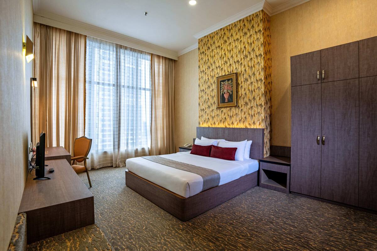 A hotel room with a large bed, a desk, a chair, a wardrobe, and a window with curtains.