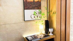 Une œuvre d’art encadrée représentant un paysage urbain, avec une vue sur les grat-ciel de la ville et un texte, un meuble en bois, un vase avec des plantes vertes, un chandelier blanc et une table avec une lampe et des magazines.