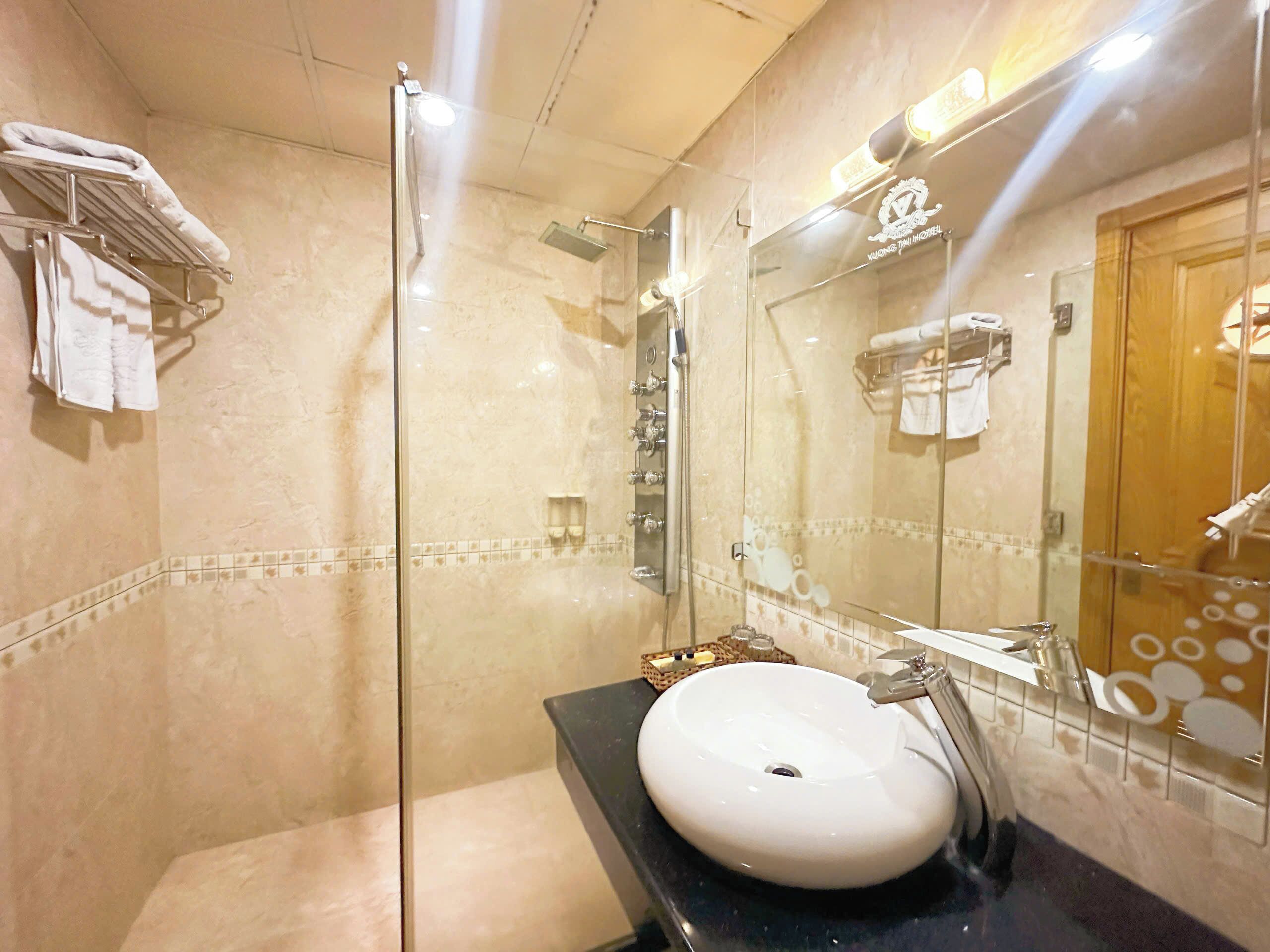 A bathroom with a glass-enclosed shower, a white sink, and a mirror reflecting a towel rack and a wooden door.