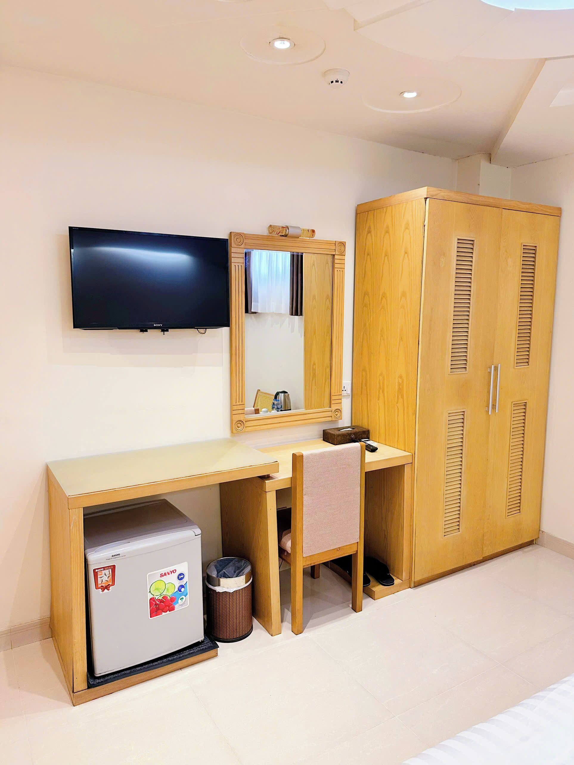 Una habitación con un escritorio de madera, un televisor montado en la pared, un armario y un refrigerador pequeño.