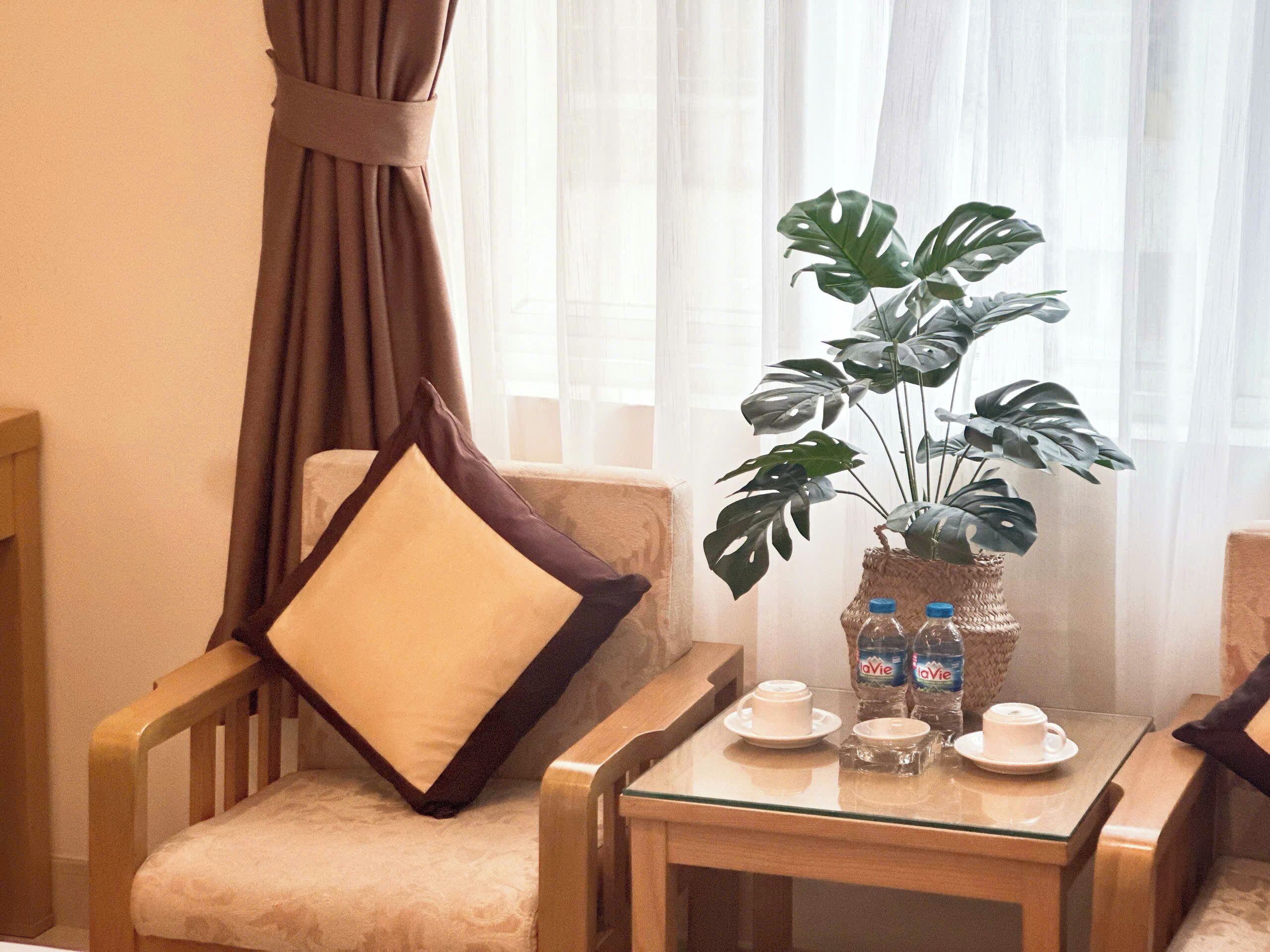 Une chambre d’hôtel avec une table en verre sur laquelle sont disposés des tasses et des soucoupes, une plante en pot et une chaise avec un coussin.
