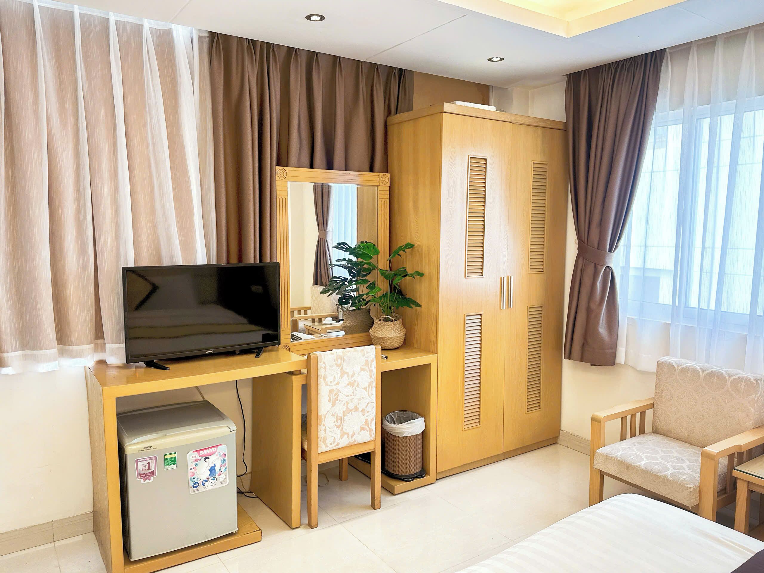 A hotel room with a wooden desk, a television, a small refrigerator, a waste bin, a potted plant, a wardrobe, and a sofa.