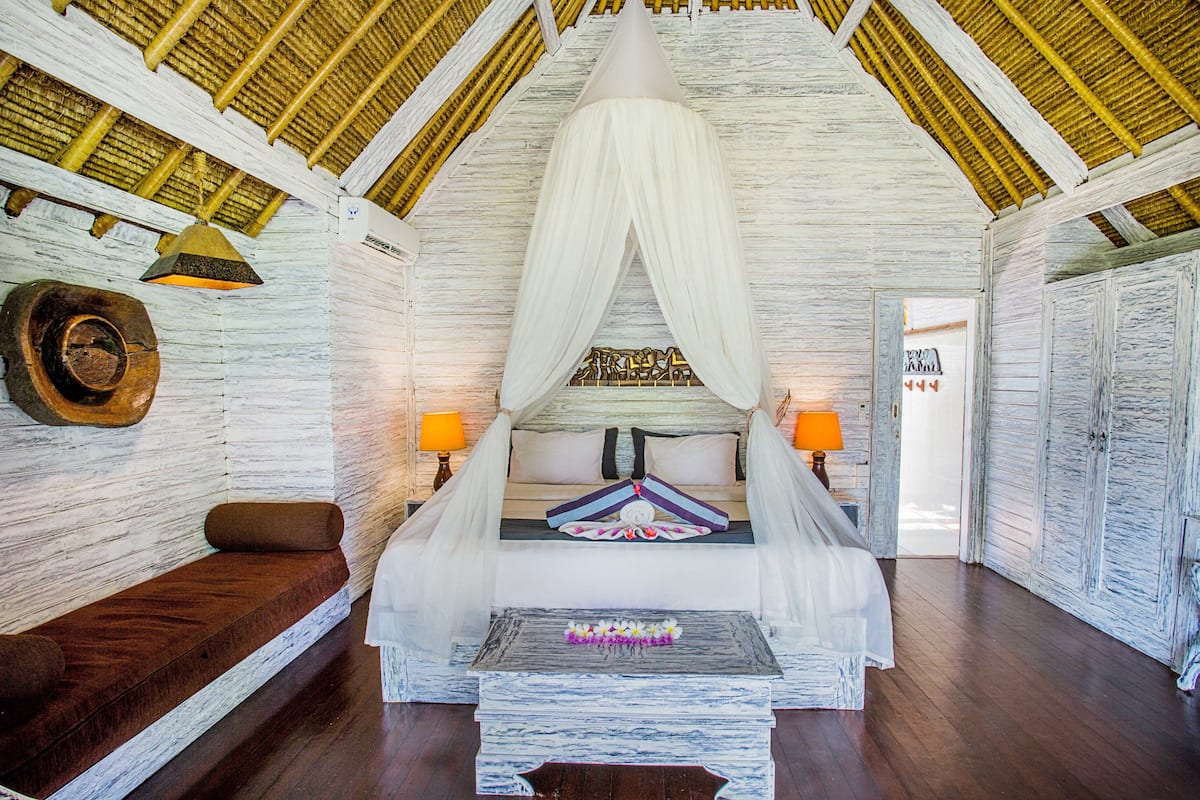 Una habitación con una cama bajo un dosel blanco, un banco de madera, una mesita con un adorno y una puerta con un letrero.