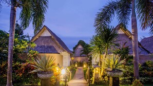 A pathway leading to a row of thatched-roof buildings, surrounded by tropical plants and palm trees.