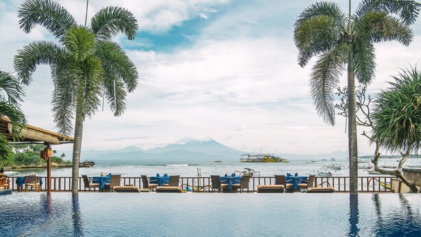 A poolside area with lounge chairs and tables, a view of the sea, and a mountain in the background.