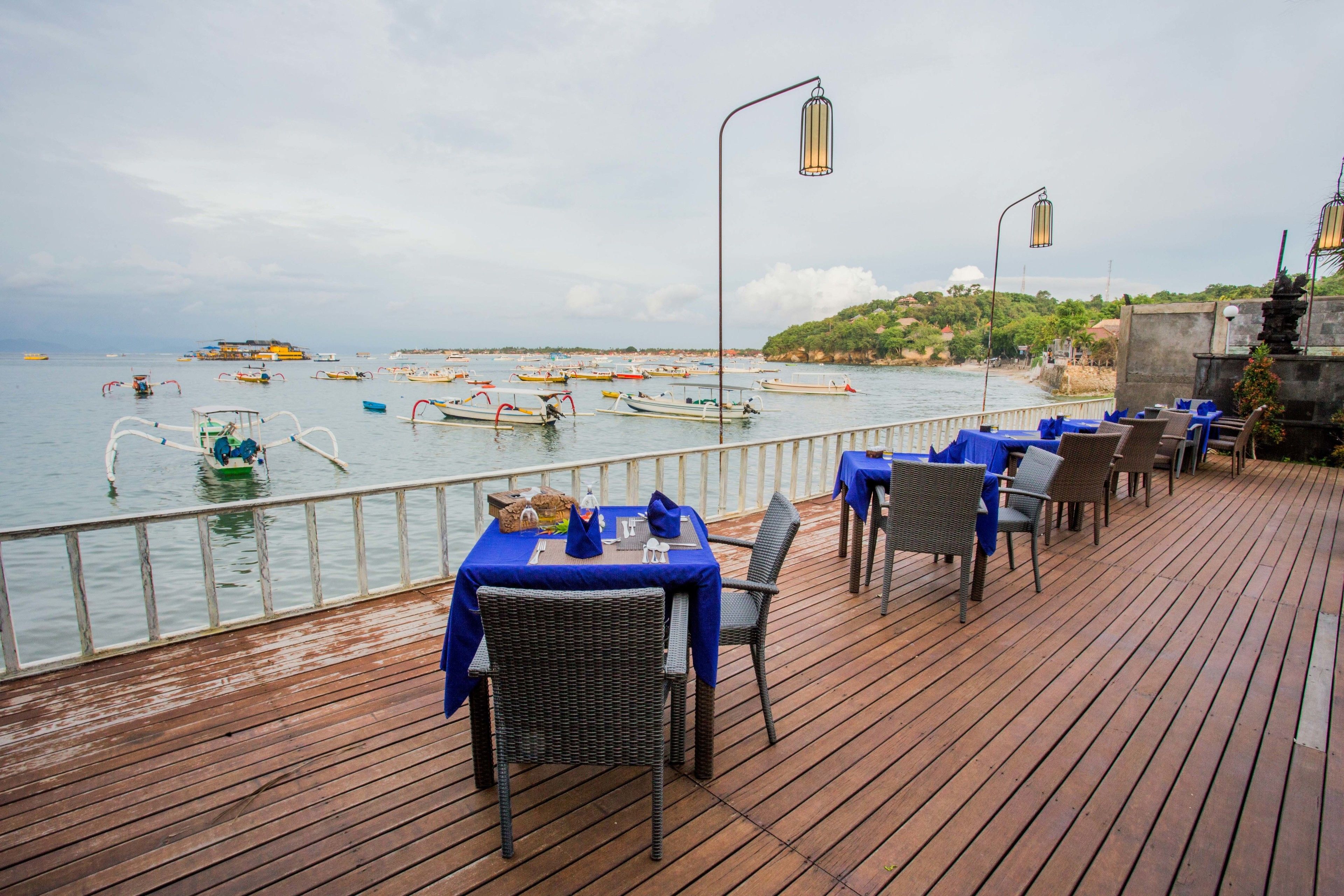 Una terraza de madera con mesas y sillas, con vista a un puerto deportivo con barcos.