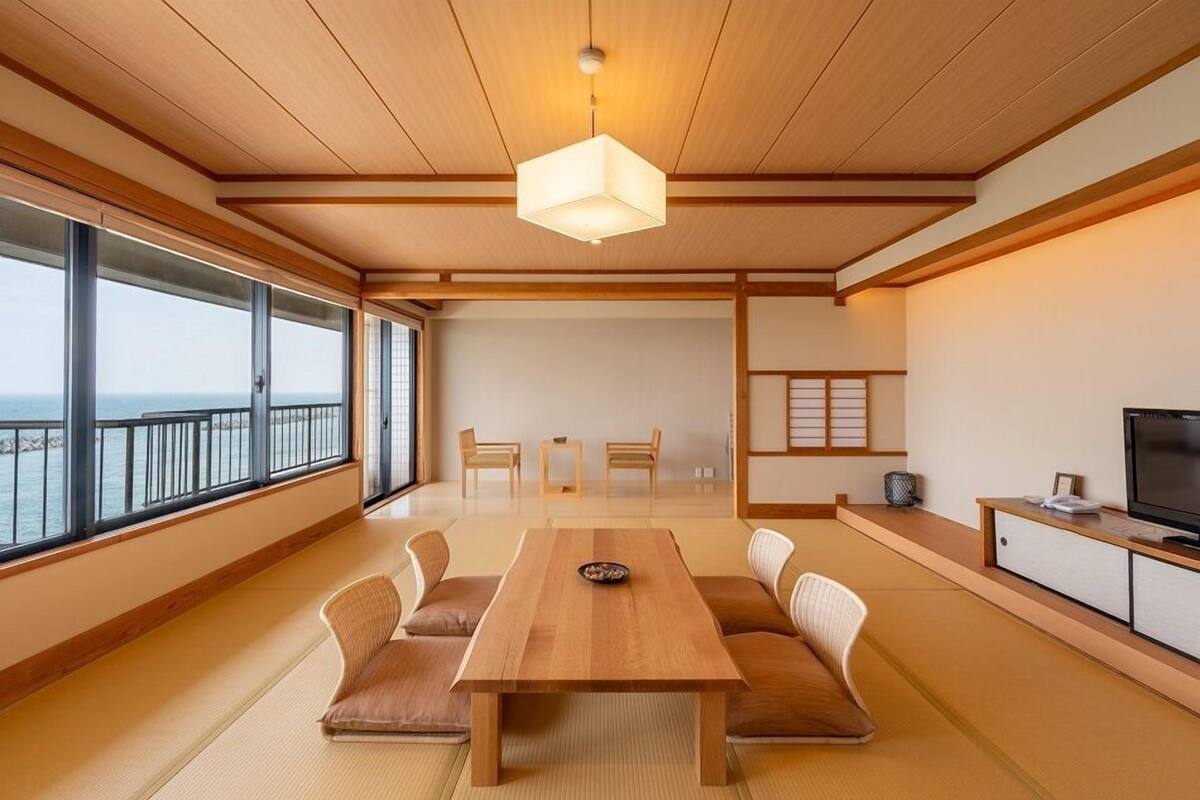 Een traditionele Japanse kamer met tatami vloeren, een lage houten tafel, tatami zitplaatsen, een televisie en grote ramen met uitzicht op zee.